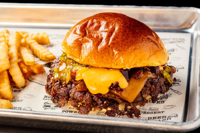 Fotografia de hamburguesa con carne doble smash, queso cheddar, bacon caramelízado y mayonesa, con patatas fritas.